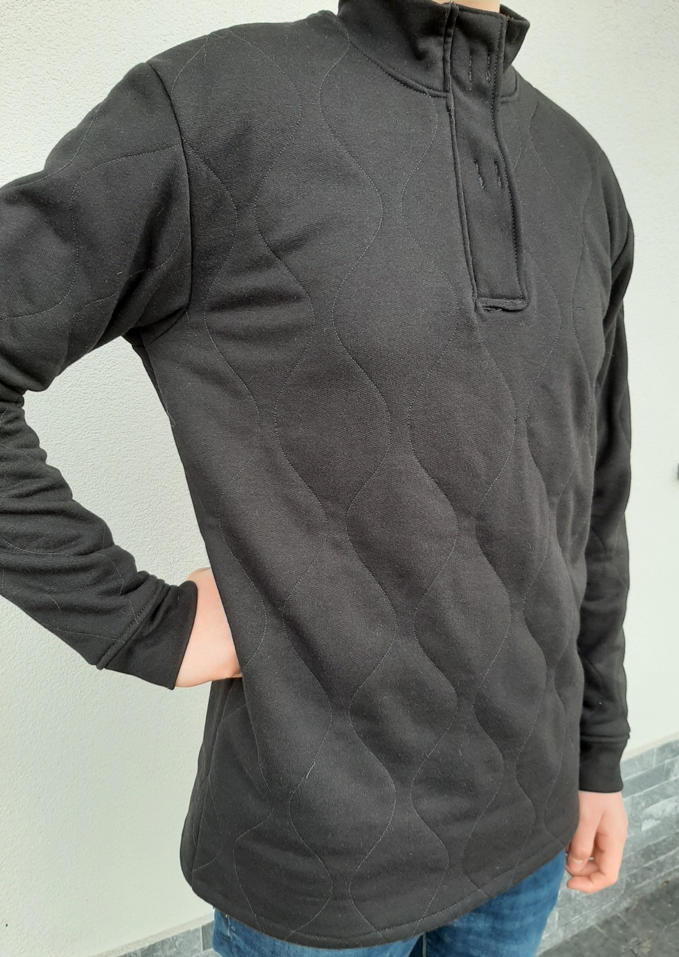 Osoba w czarnej, pikowanej bluzie z krótkim zapięciem na trzy guziki, stojąca na tle jasnej ściany.