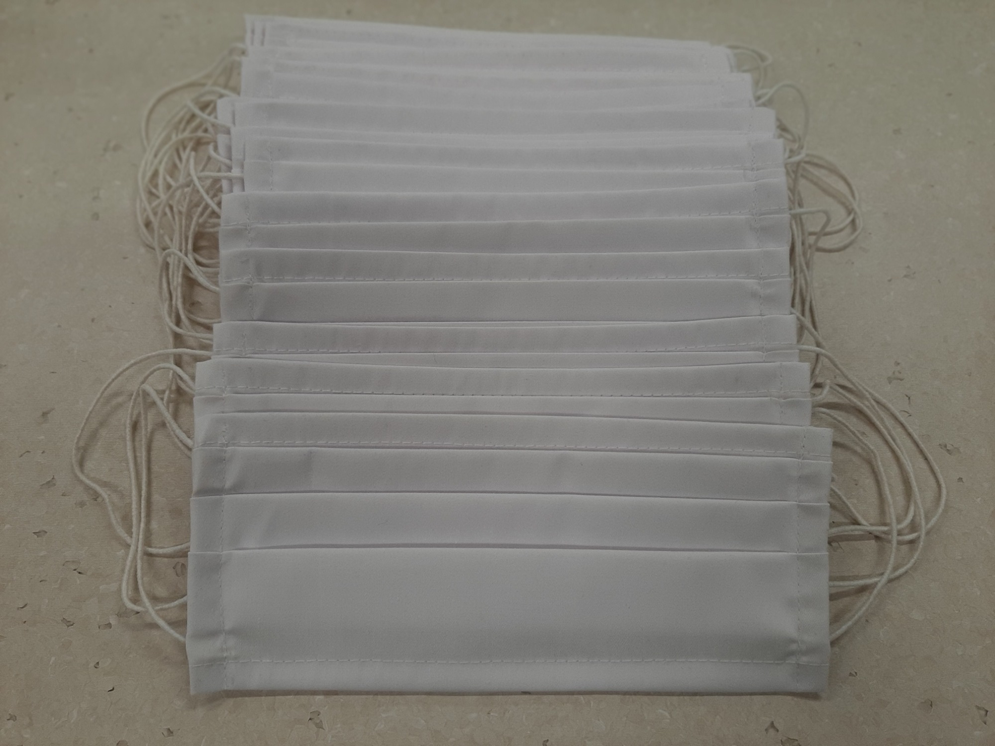 Stos złożonych, białych, plisowanych maseczek ochronnych z gumkami, ułożonych na beżowym blacie.