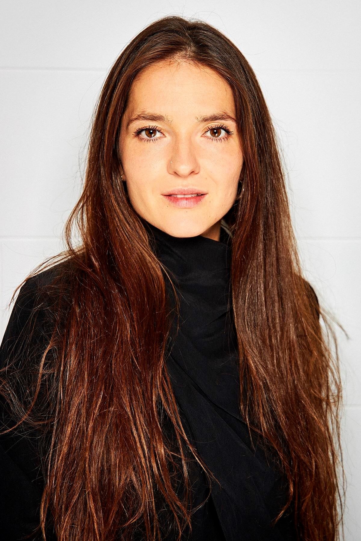 Portret kobiety z długimi, brązowymi włosami i delikatnym makijażem, ubrana w czarny golf, na jasnym tle.