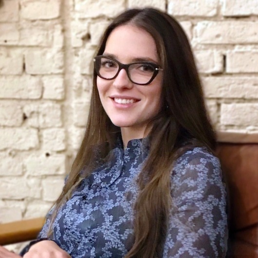 Uśmiechnięta kobieta w okularach o kocim kształcie i kwiecistej bluzce na tle ceglanej ściany.