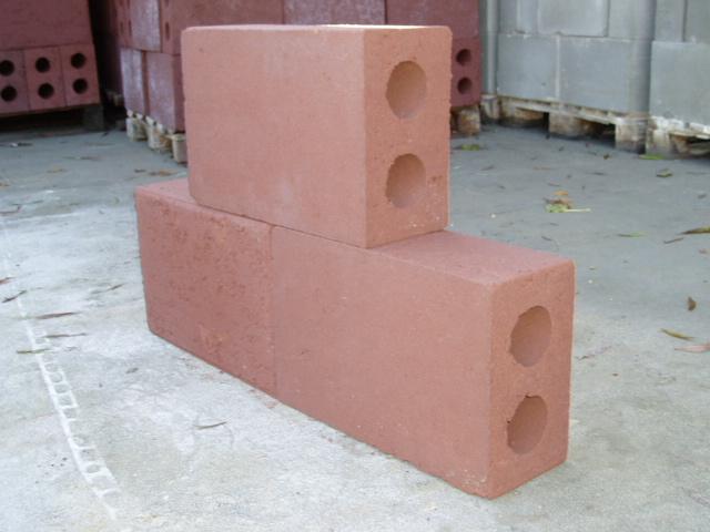 Dwa bloczki keramzytobetonowe w kolorze ceglastym ułożone jeden na drugim, z dwoma okrągłymi otworami w każdym, na betonowej posadzce, z paletą bloczków w tle.