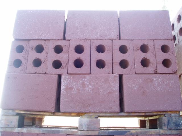 Ułożone na palecie bloczki keramzytobetonowe w odcieniach czerwieni, niektóre z czterema otworami, widoczne ślady po produkcji.
