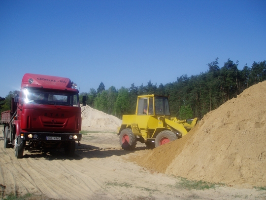 Czerwona ciężarówka marki Star i żółta ładowarka na tle hałdy piasku budowlanego i lasu w słoneczny dzień.