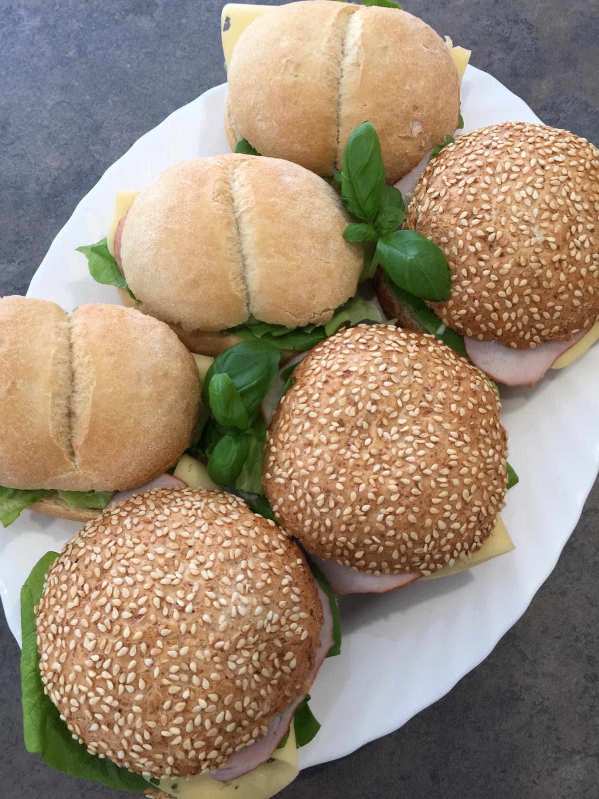 Biały talerz z sześcioma kanapkami: trzy z bułkami posypanymi sezamem i trzy z gładkimi bułkami, wszystkie z szynką, serem i sałatą, udekorowane listkami bazylii.