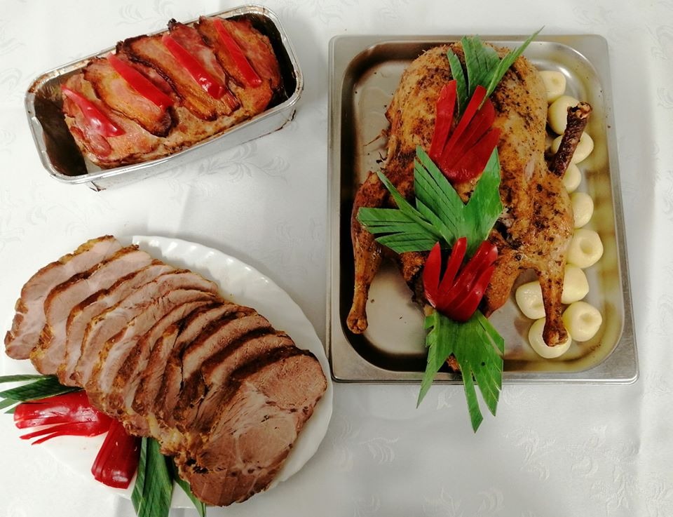 Uroczysty stół z pieczoną kaczką udekorowaną papryką i zielonymi liśćmi, pasztetem w foremce aluminiowej i plastrami pieczeni na białym talerzu, całość ozdobiona papryką i zielenią.