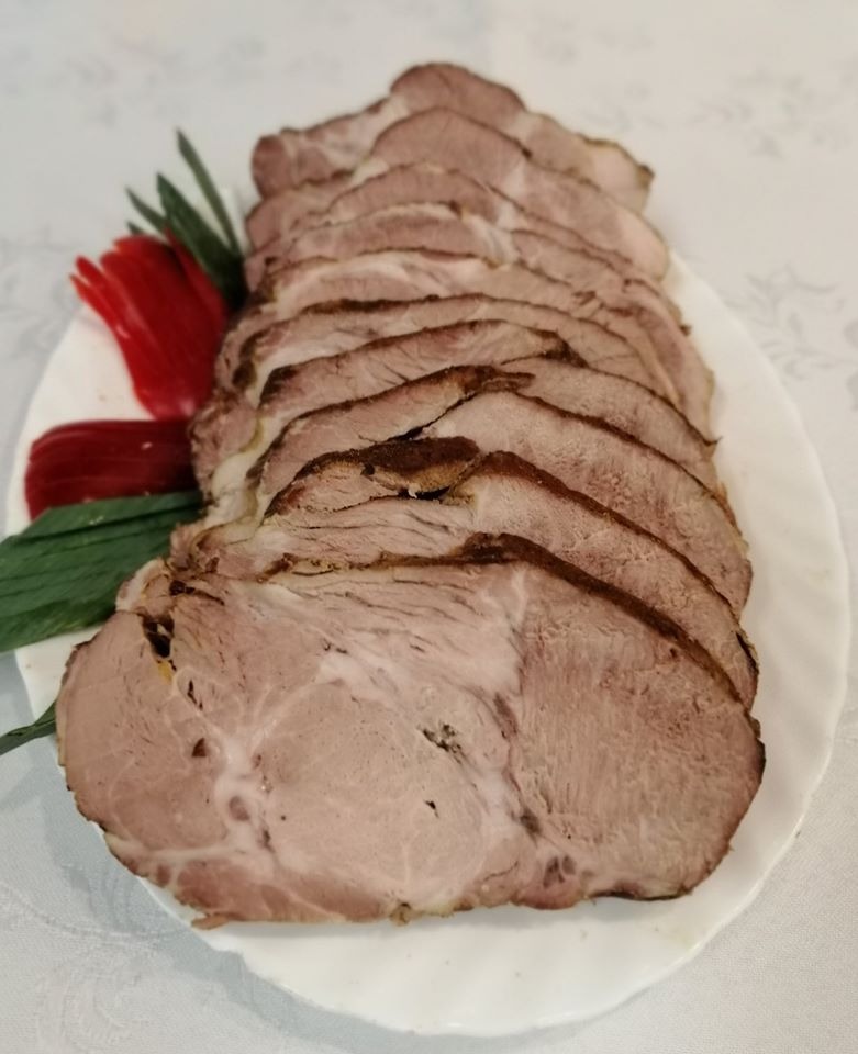 Plasterki pieczonego mięsa ułożone na białym talerzu, udekorowane czerwoną papryką i zielonymi pędami, na tle białego obrusu z delikatnym wzorem.