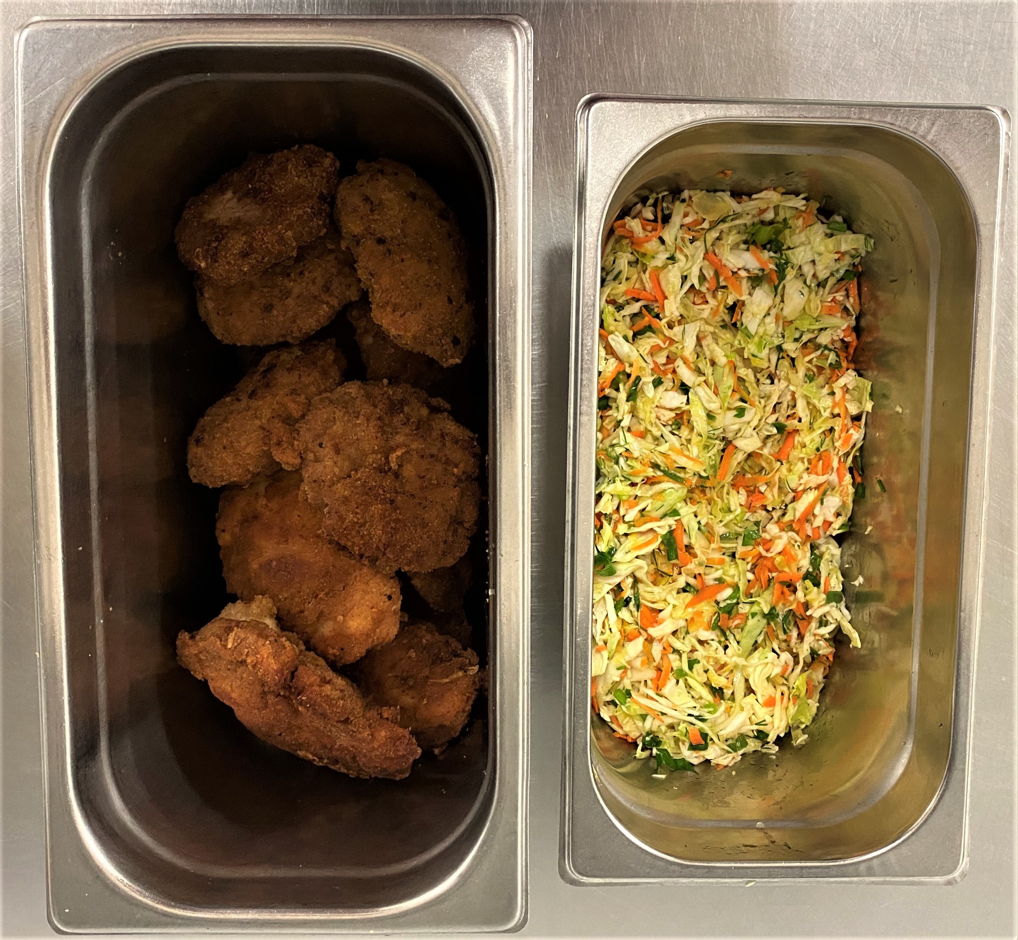 Dwa metalowe pojemniki: jeden z panierowanymi kawałkami kurczaka, drugi z surówką z kapusty i marchewki z dodatkiem zieleniny.