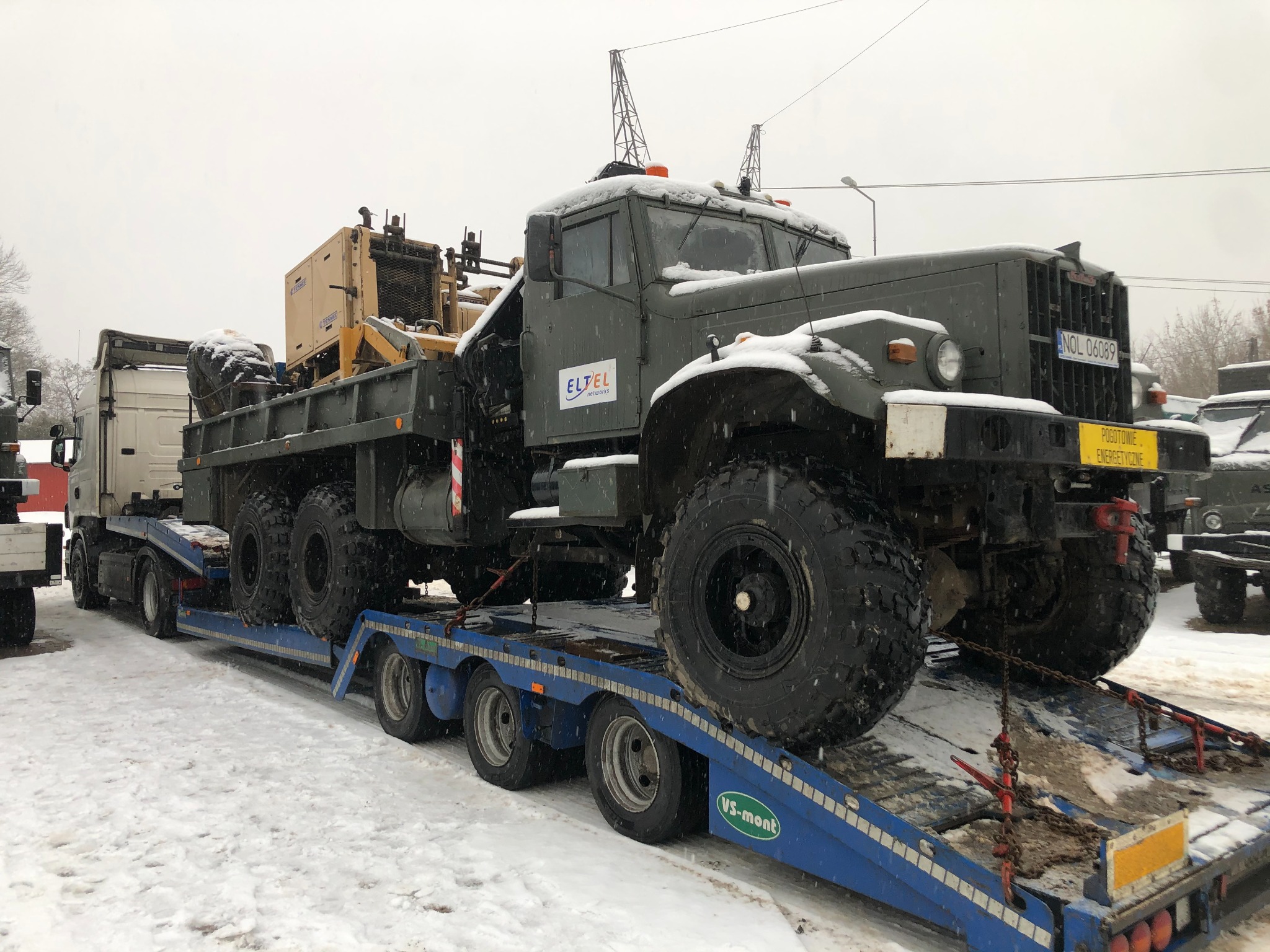 Ciężki, wojskowy samochód transportowy z logo ELTEL na lawecie w zimowej scenerii, przewożony w ramach pogotowia energetycznego.
