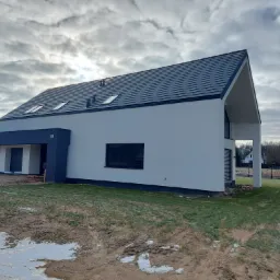 Nowoczesny dom jednorodzinny z białymi ścianami i grafitowym podcieniem, ciemna dach&oacute;wka, okna dachowe, posesja w trakcie zagospodarowywania.