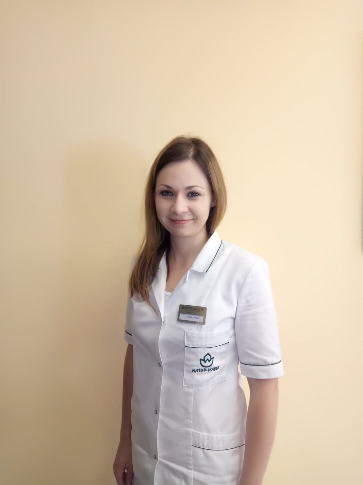 Uśmiechnięta kobieta w białym uniformie z plakietką i logo Natur House na tle beżowej ściany.