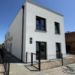 Nowoczesny, biały dom z czarnymi ramami okiennymi i numerami 69 i 71 na elewacji, otoczony czarnym ogrodzeniem panelowym na tle błękitnego nieba.