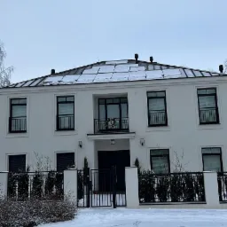 Dwukondygnacyjny dom w stylu rezydencjalnym z płaskim dachem pokrytym śniegiem, czarnymi ramami okiennymi i balkonem z dekoracją kwiatową, otoczony zimowym krajobrazem.