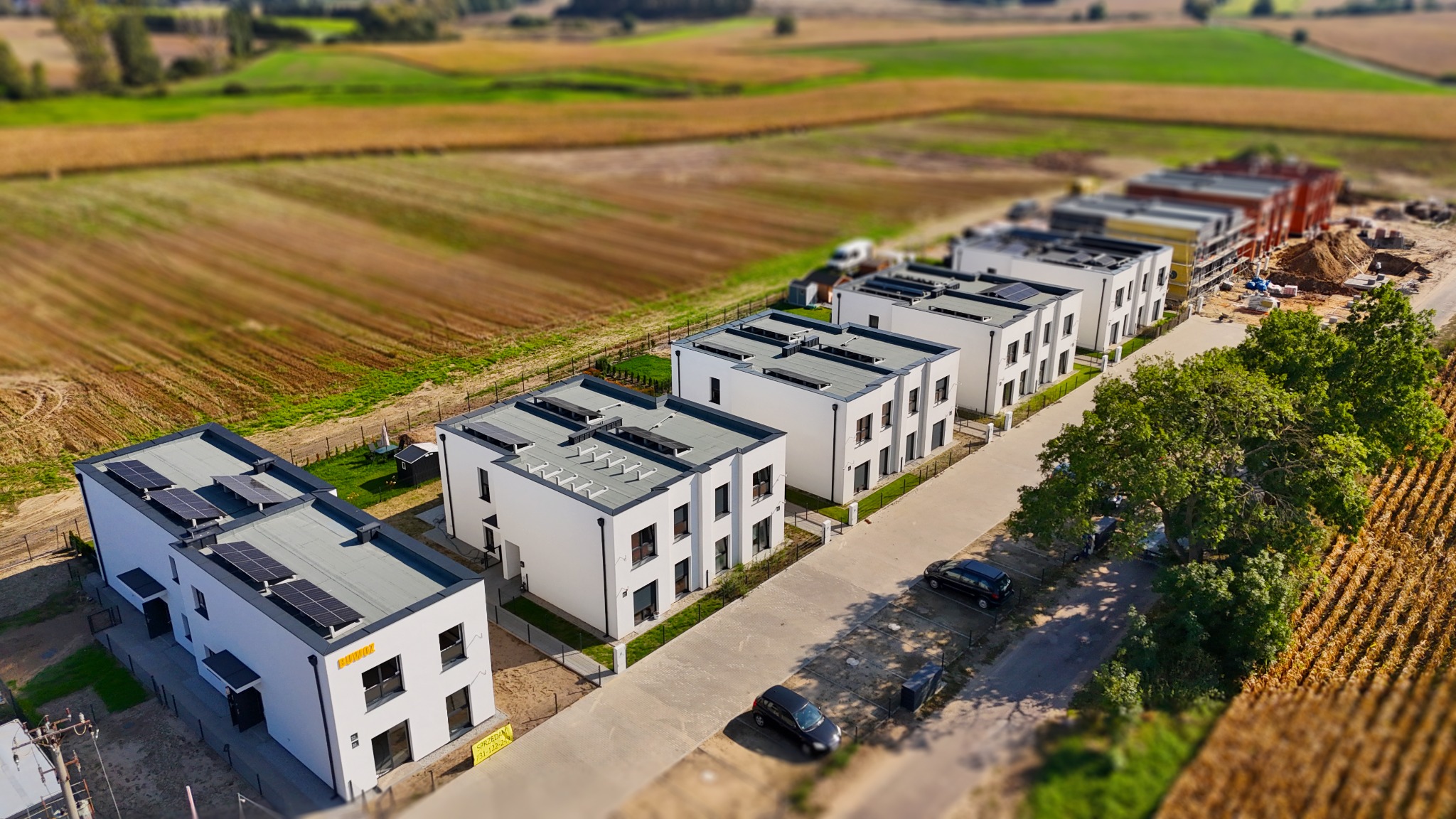 Nowoczesne, białe domy szeregowe z panelami słonecznymi na dachu, widok z lotu ptaka, w tle pola uprawne i domy w trakcie budowy.
