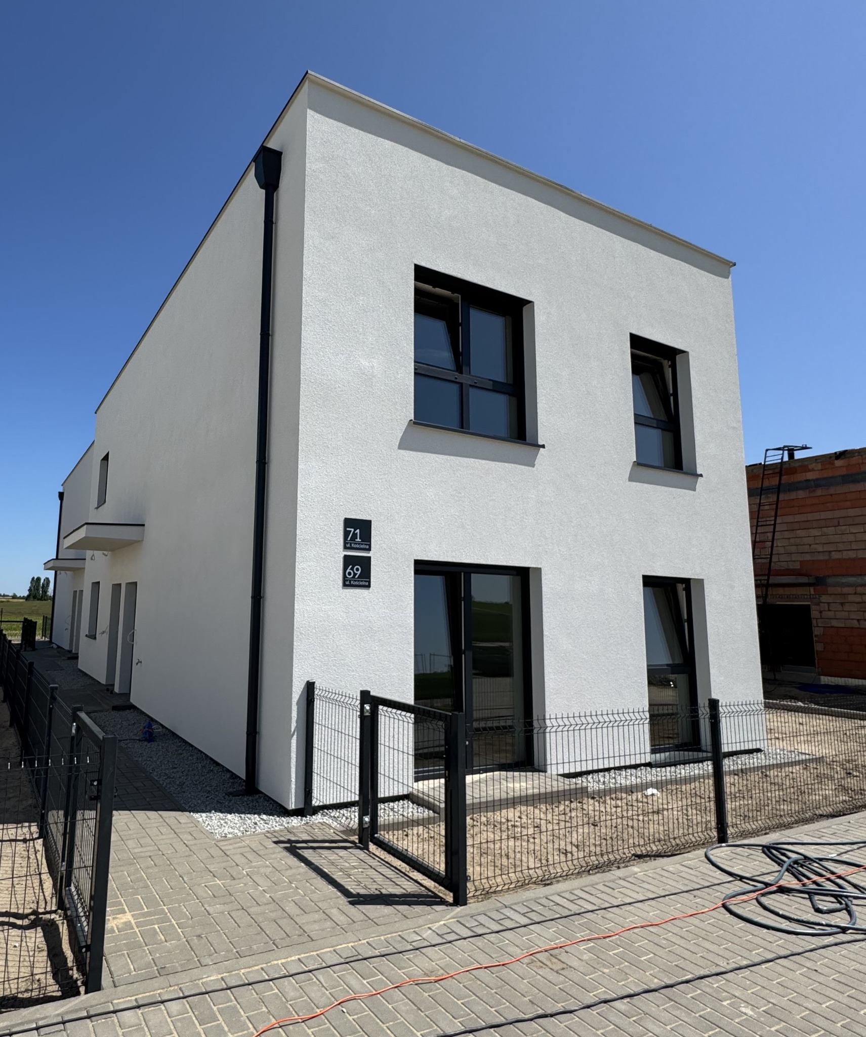 Nowoczesny, biały dom z czarnymi ramami okiennymi i numerami 69 i 71 na elewacji, otoczony czarnym ogrodzeniem panelowym na tle błękitnego nieba.