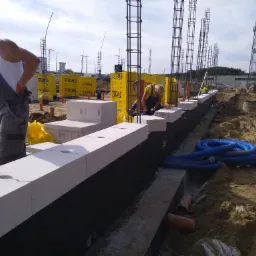 Budowa fundamentów z bloczków Ytong, widoczni dwaj pracownicy w trakcie pracy, zbrojenie pionowe, niebieskie rury na ziemi, w tle składowane bloczki Ytong na paletach.