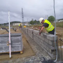 Pracownik w kasku i kamizelce odblaskowej muruje ścianę z szarych bloczków betonowych na placu budowy, widoczny poziomica laserowa, paleta z bloczkami i wiertarka akumulatorowa.