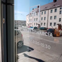 Widok przez szybę biura na ulicę z zaparkowanymi samochodami, na szybie naklejone logo firmy i godziny otwarcia punktu.