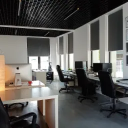 Jasne biuro z rzędami biurek, krzeseł obrotowych i komputerów Apple, ozdobione certyfikatami na ścianie i lampą z abażurem z papieru ryżowego.