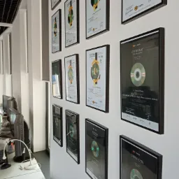 Ściana biura z powieszonymi certyfikatami ukończenia szkoleń IT Media, prezentującymi nazwiska uczestników i nazwy programów, w czarnych ramkach.