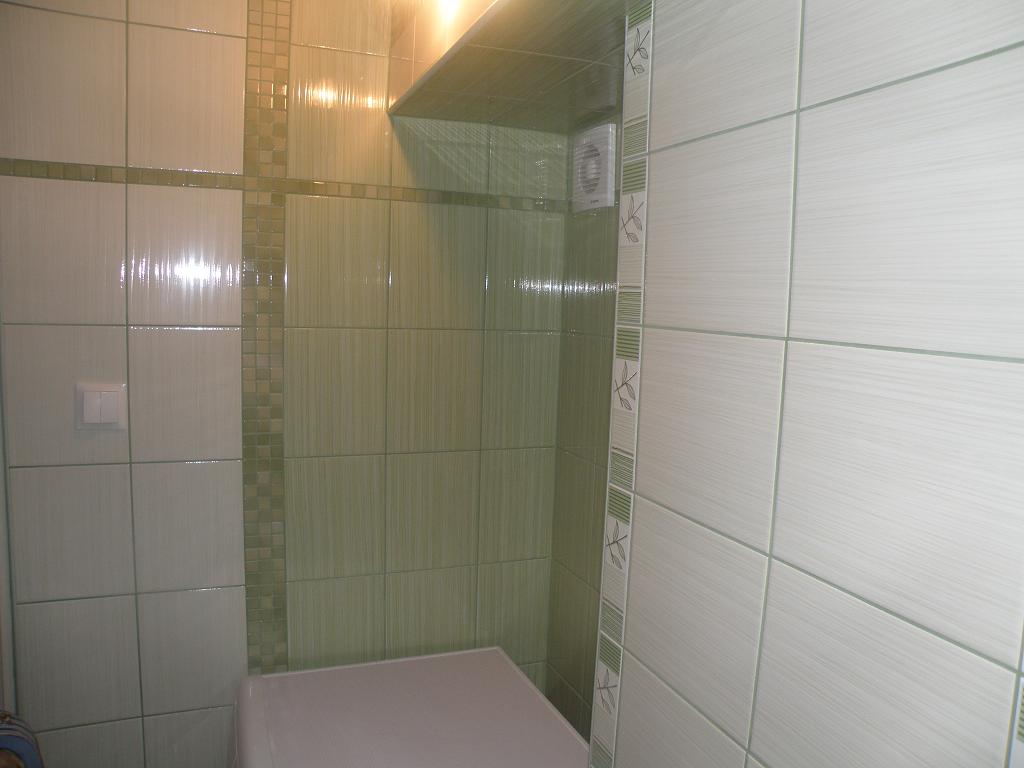 Wykończona łazienka z jasnymi, kwadratowymi płytkami w odcieniach zieleni, z dekoracyjnymi paskami płytek mozaikowych i z motywem liści, widoczny włącznik światła i kratka wentylacyjna.