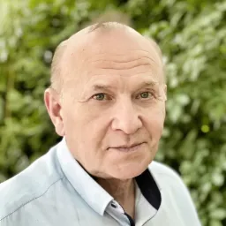 Portret starszego mężczyzny w błękitnej koszuli na tle zieleni, z delikatnym uśmiechem i zmarszczkami wokół oczu.