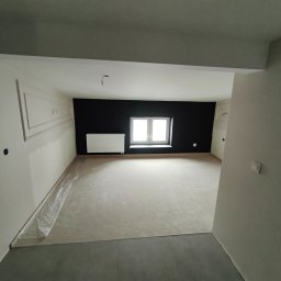 SANY-BUD SPÓŁKA Z OGRANICZONĄ ODPOWIEDZIALNOŚCIĄ - Wnętrze po wylewce, ściany częściowo pomalowane na biało, jedna ściana czarna, okno z grzejnikiem. Widoczne listwy dekoracyjne na ścianie.