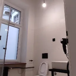 Minimalistyczna łazienka z ceglanym sufitem, wiszącą toaletą i umywalką na metalowym stelażu, oświetlona pojedynczymi żarówkami.