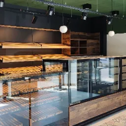 Nowoczesne wnętrze piekarni-cukierni z drewnianymi regałami, szklanymi witrynami chłodniczymi i dekoracyjną ścianą z mozaiką.