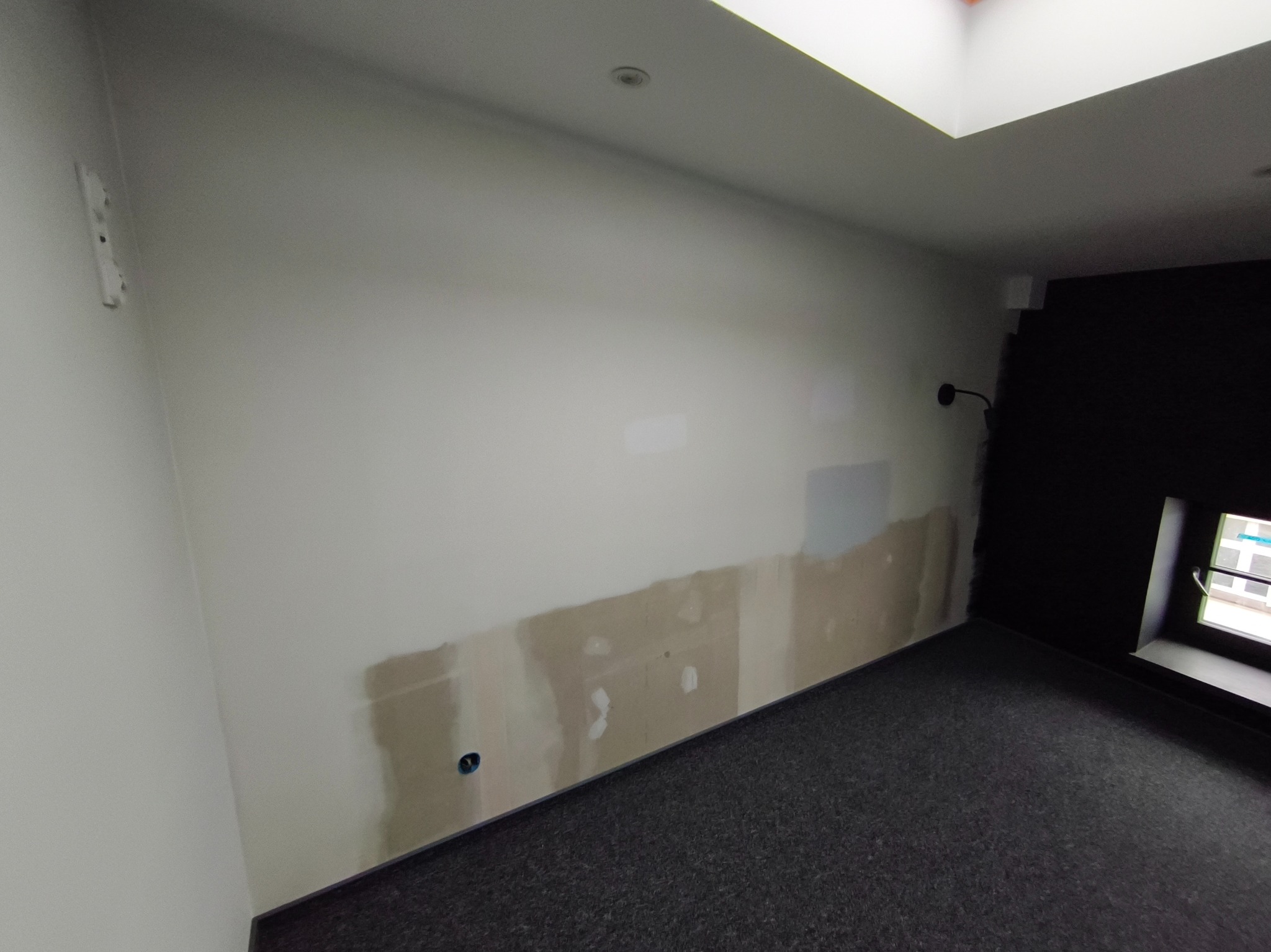 Ściana w trakcie szpachlowania z widocznymi łatami i gładzią, przygotowywana do malowania. Fragment podłogi z ciemną wykładziną. Okno z czarną ramą.