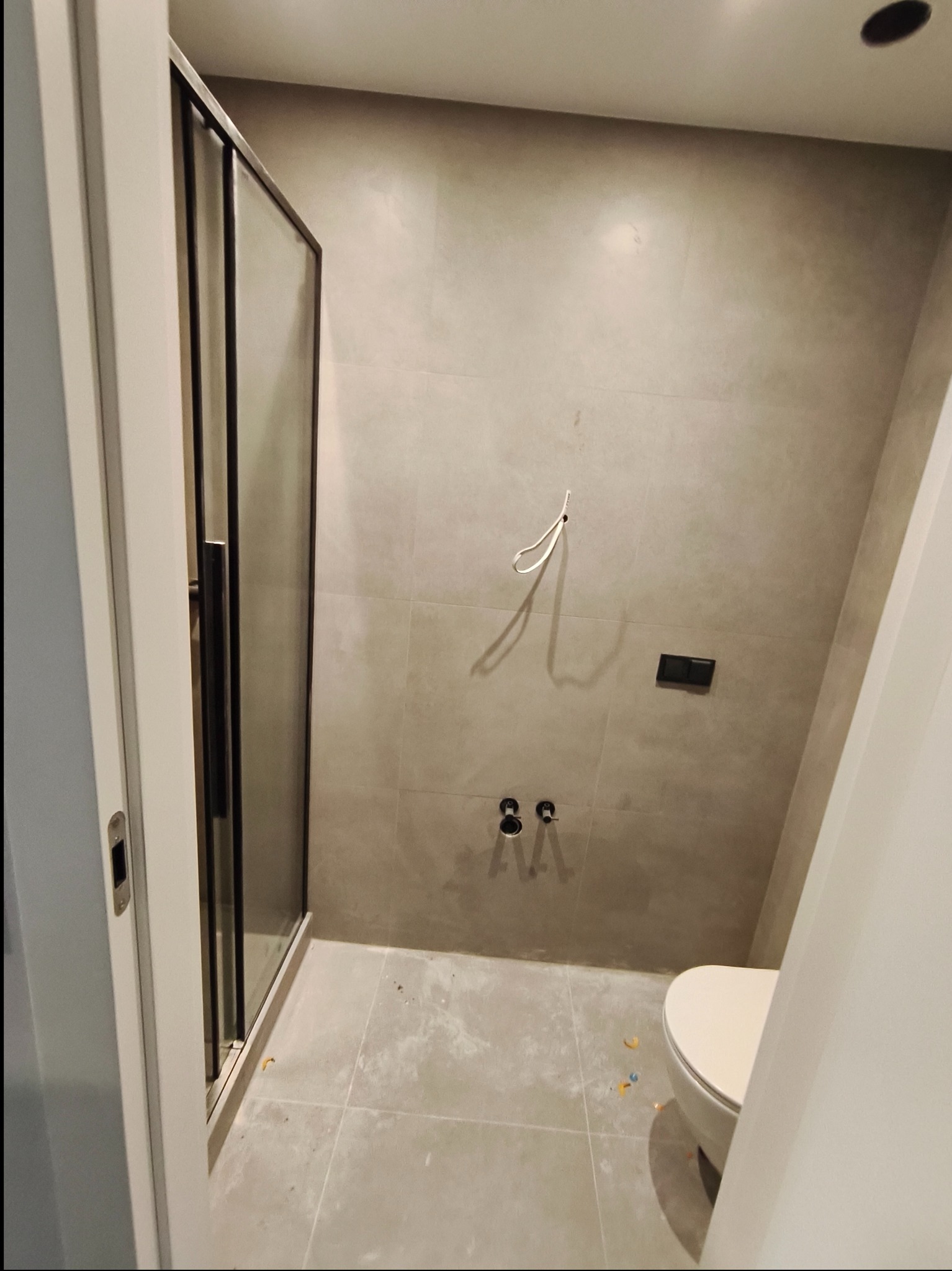 Wykończona łazienka z kabiną prysznicową z czarnymi profilami, szarą glazurą i wiszącą toaletą. Widoczne przyłącza wodne i fragment instalacji elektrycznej.