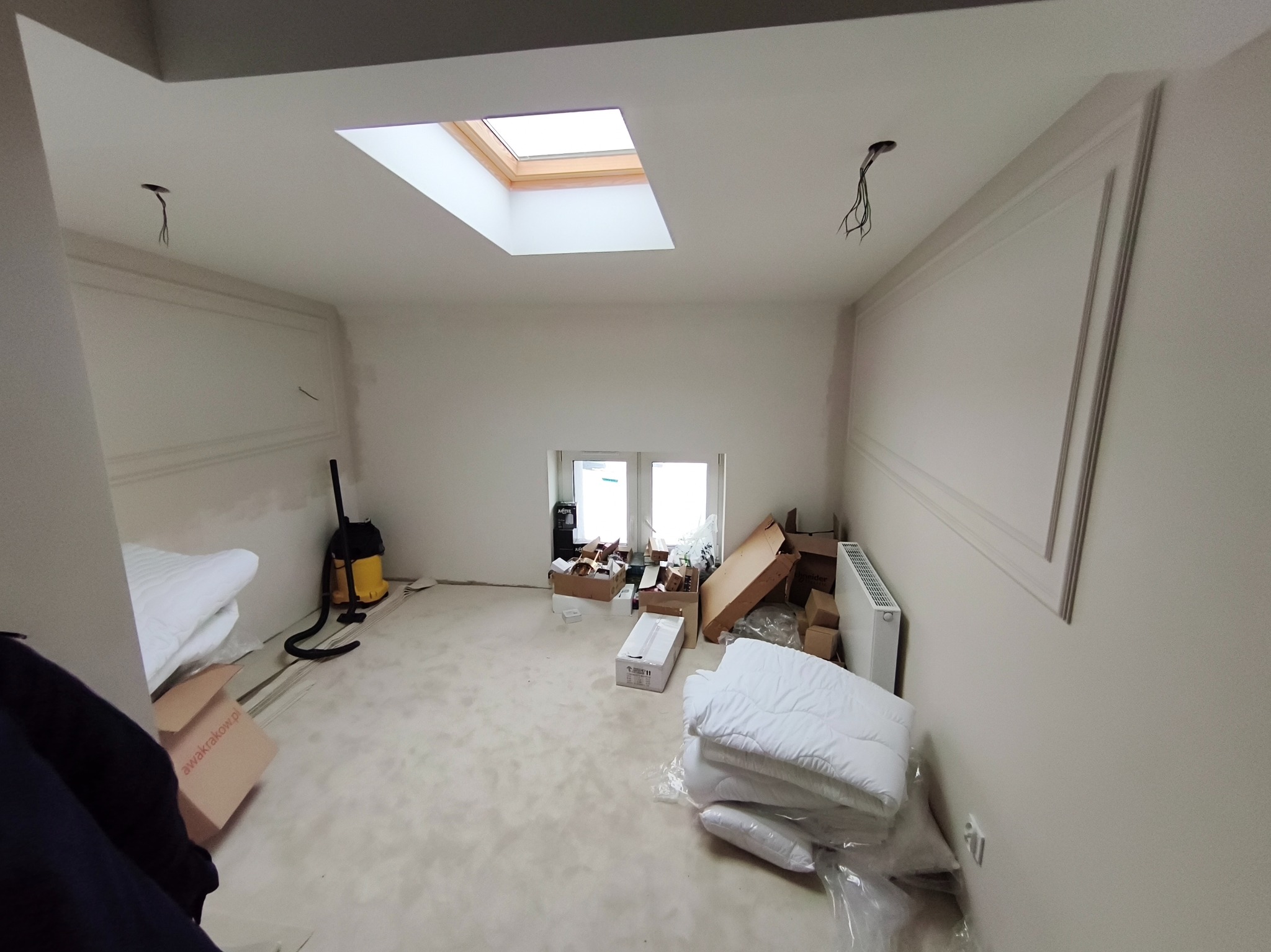 Pokój w trakcie wykańczania: widoczne listwy dekoracyjne na ścianach, okno dachowe i okno na wprost. Na podłodze kartony i narzędzia, sugerujące trwające prace wykończeniowe.