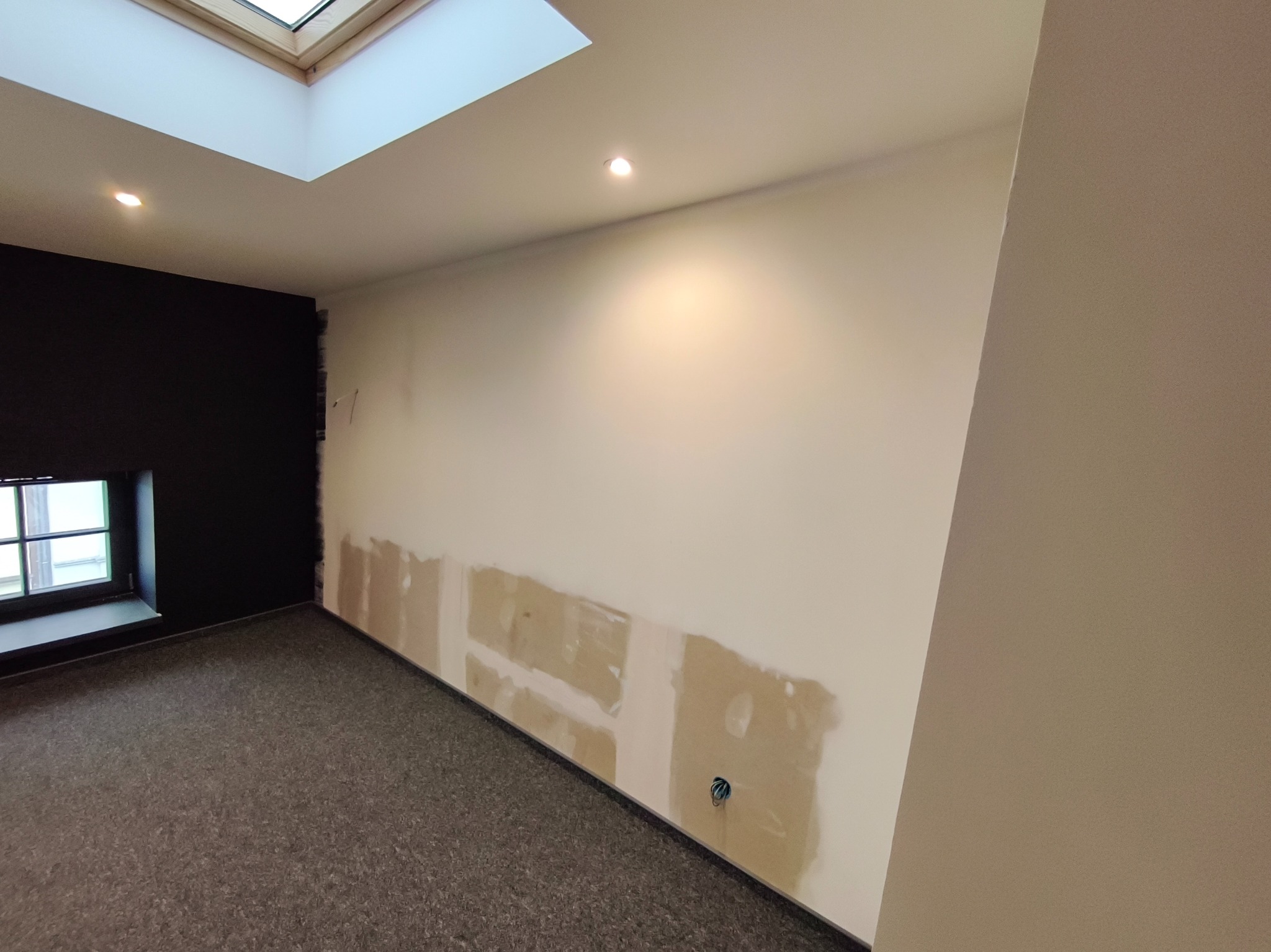 Wnętrze w trakcie remontu: fragment ściany z widocznymi śladami szpachlowania, okno z czarną ramą i roletą, oraz sufit z oknem dachowym i punktowym oświetleniem.