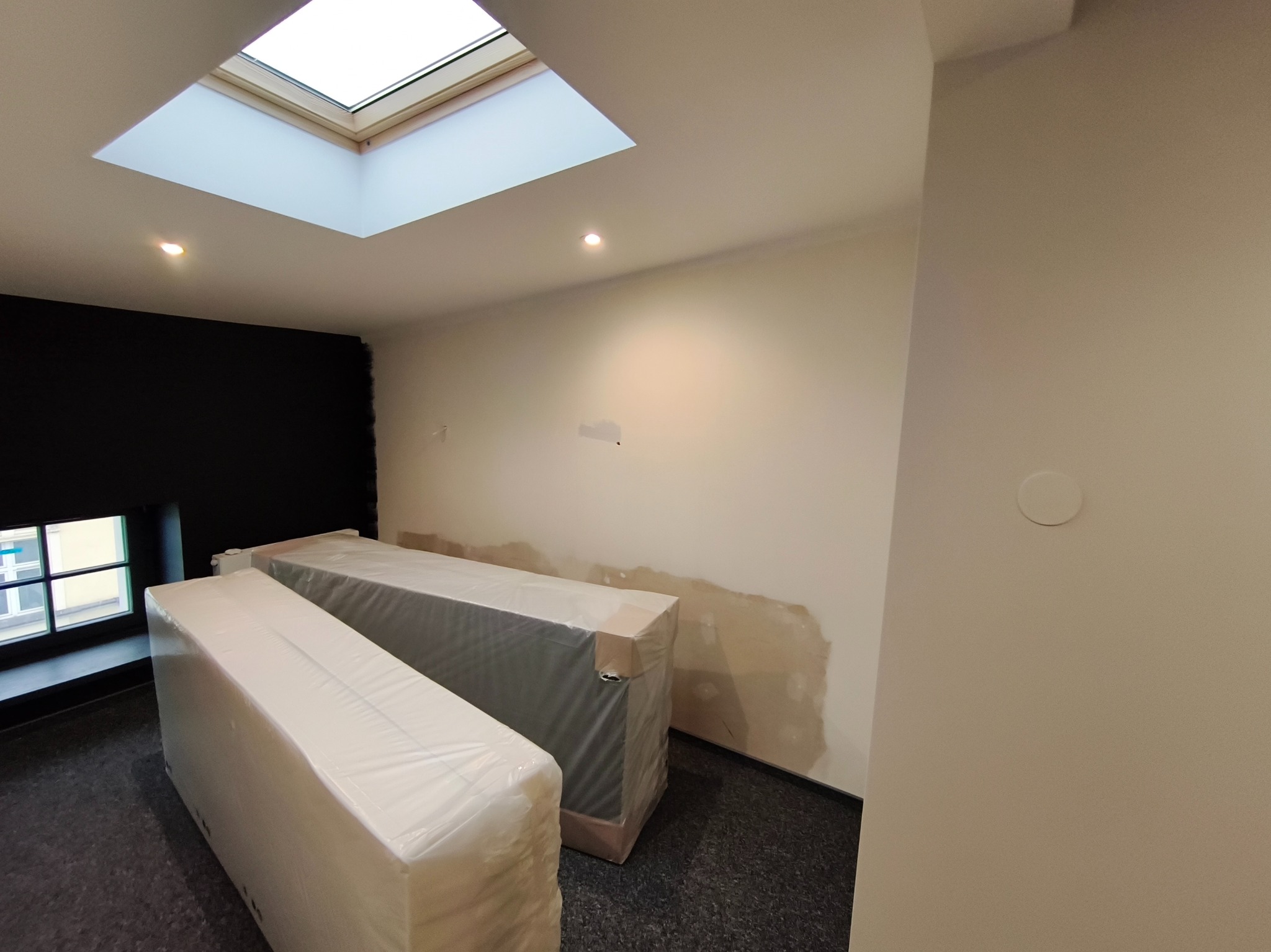 Wnętrze w trakcie wykańczania: ściany przygotowane do malowania, dwa meble zabezpieczone folią, okno dachowe w suficie. Widoczne ślady po szpachlowaniu.