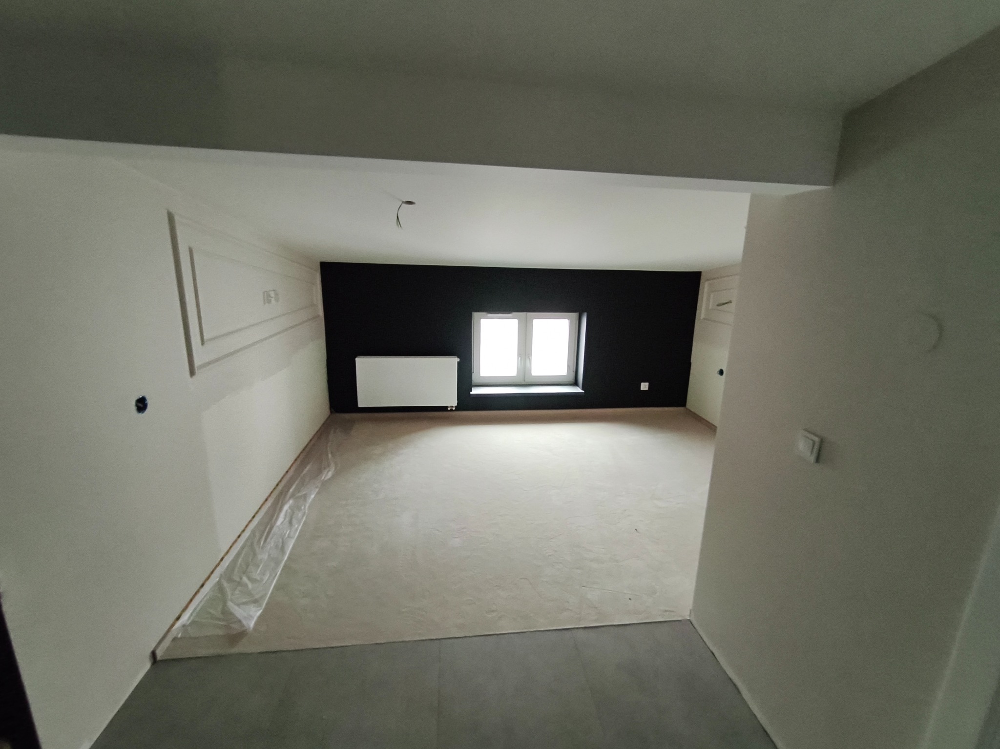 Wnętrze po wylewce, ściany częściowo pomalowane na biało, jedna ściana czarna, okno z grzejnikiem. Widoczne listwy dekoracyjne na ścianie.