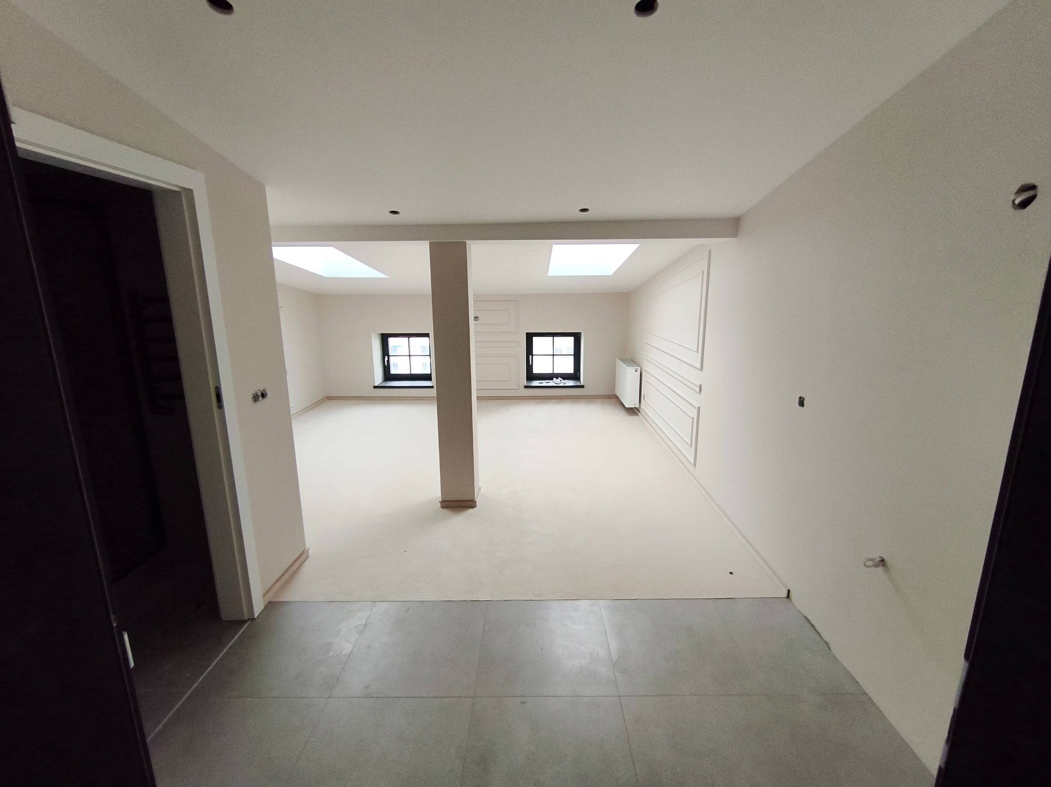 Jasne, przestronne wnętrze po remoncie z kolumną, oknami dachowymi i dekoracyjną sztukaterią na ścianie. Widok z korytarza na świeżo wykończone pomieszczenie.