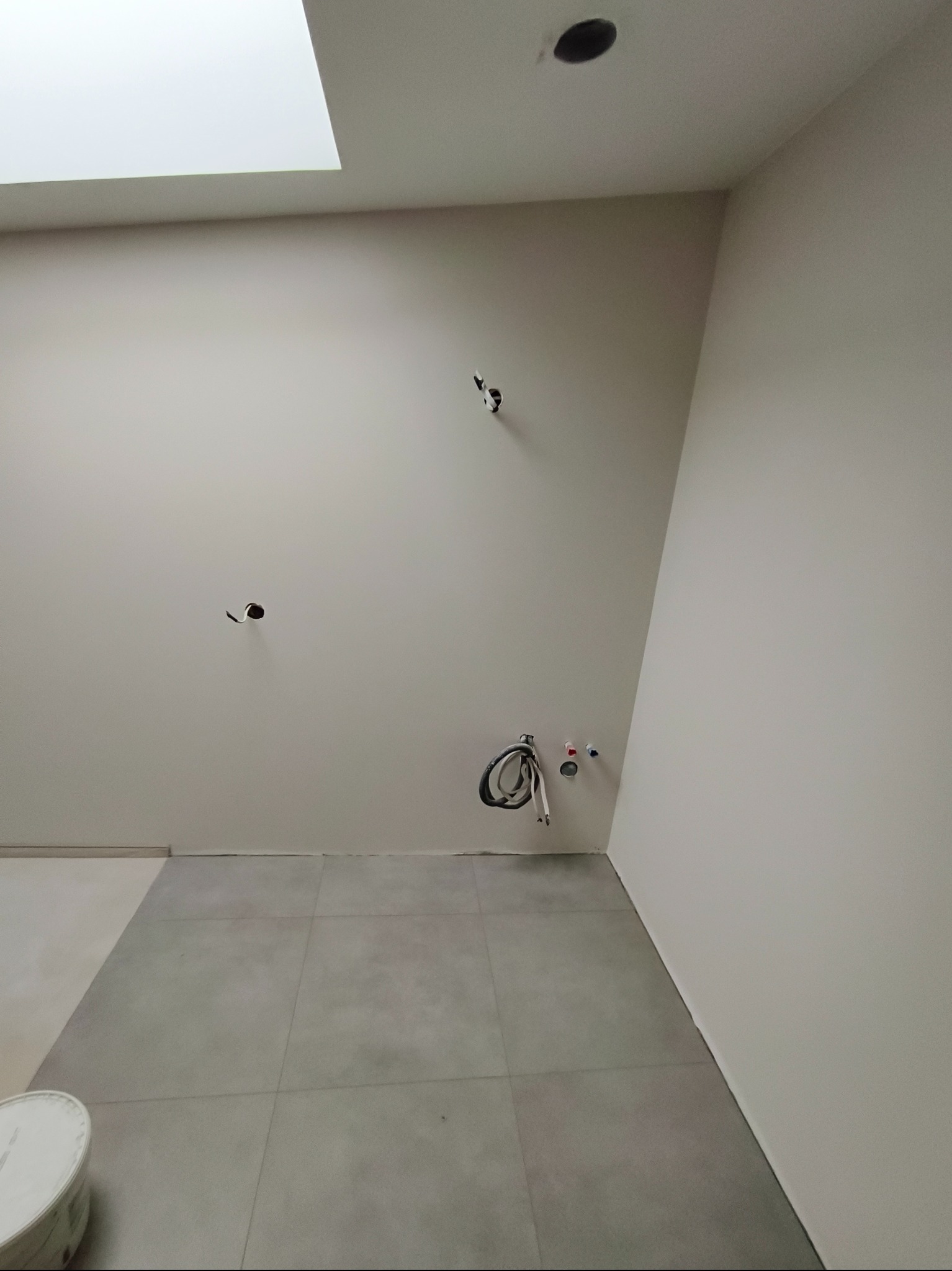 Jasna łazienka w trakcie wykańczania: szare płytki podłogowe, gładkie ściany z wyprowadzonymi instalacjami, okno dachowe. Minimalistyczny design w neutralnej kolorystyce.