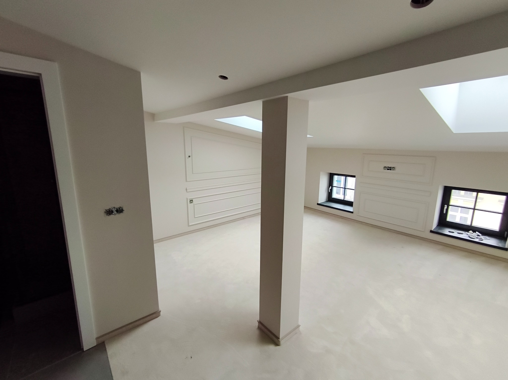 Jasne, przestronne wnętrze po remoncie z kolumną, sztukaterią na ścianach i oknami w czarnych ramach. Widoczne otwory na oświetlenie w suficie.