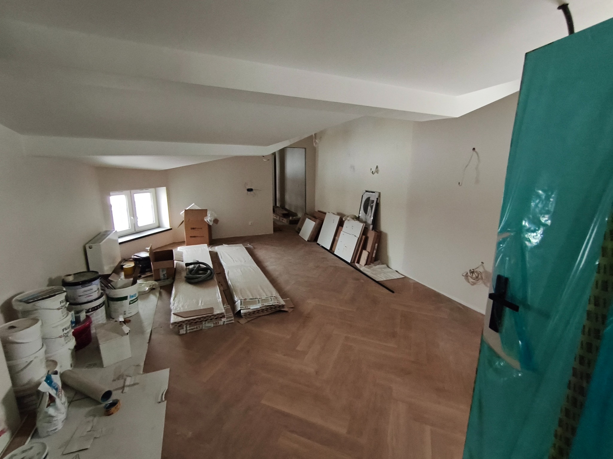 Jasne, przestronne poddasze z parkietem w jodełkę, przygotowane do dalszych prac wykończeniowych. Widoczne materiały budowlane i zabezpieczone drzwi.