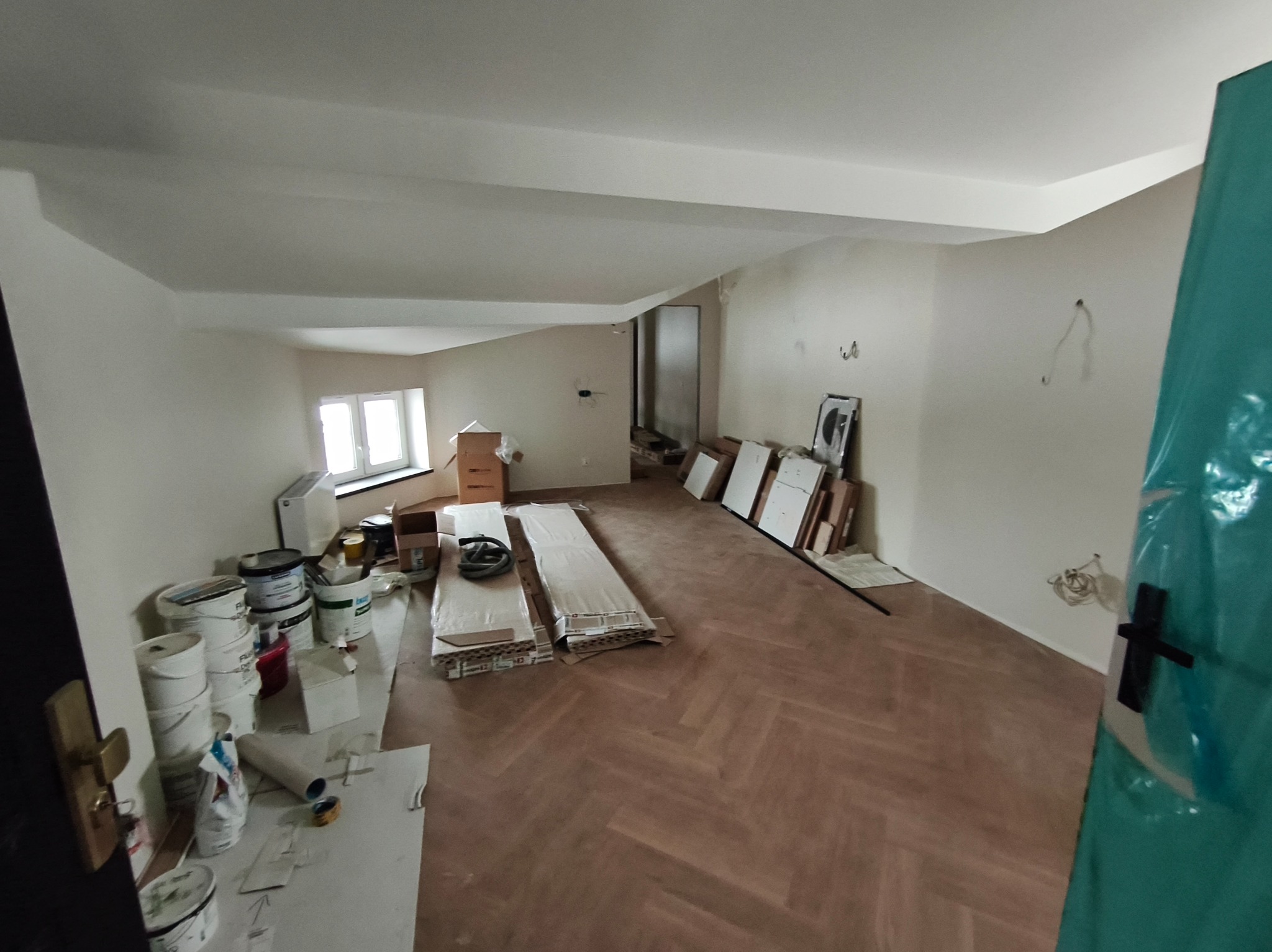Wnętrze poddasza w trakcie remontu: parkiet w jodełkę, białe ściany, okno, farby, płyty. Widok zza uchylonych drzwi zabezpieczonych folią malarską.