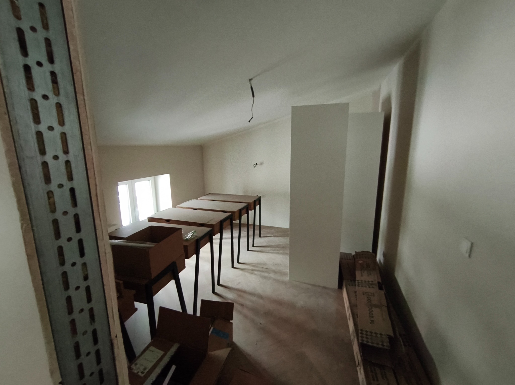 Wnętrze poddasza w trakcie wykańczania: widoczne stelaże, płyty gipsowe i kartony. Przygotowania do montażu ścianek działowych i sufitu. Oświetlenie naturalne z okna.