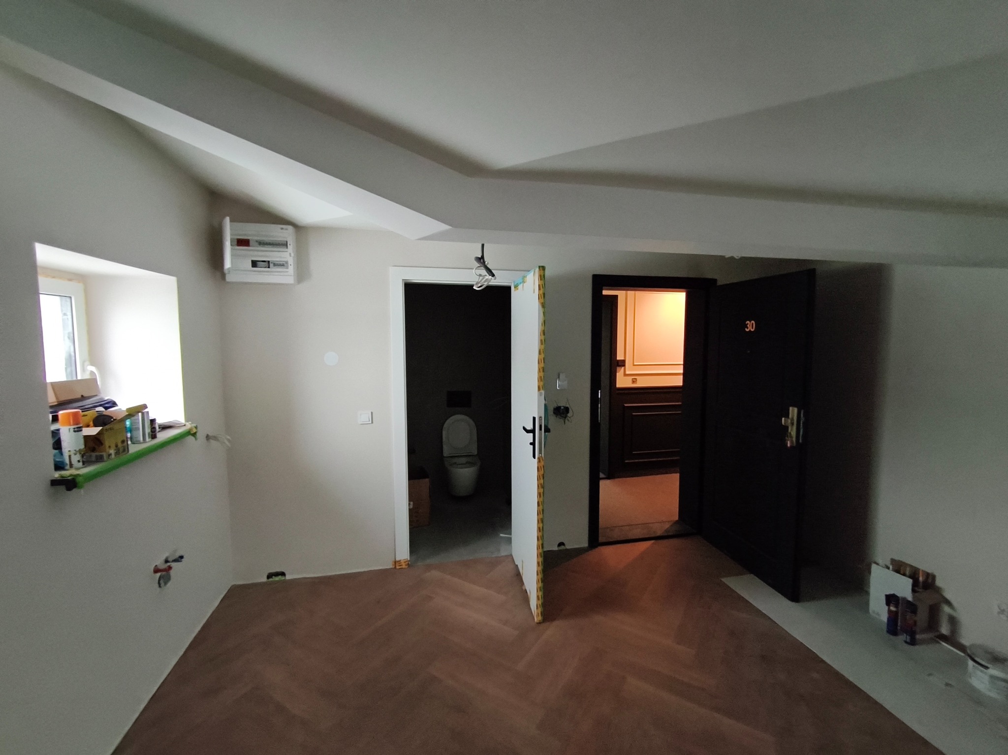 Pokój po remoncie z parkietem w jodełkę, otwartymi drzwiami do łazienki i pokoju. Widoczne puszki z farbą i narzędzia, sugerujące świeżo zakończone prace wykończeniowe.