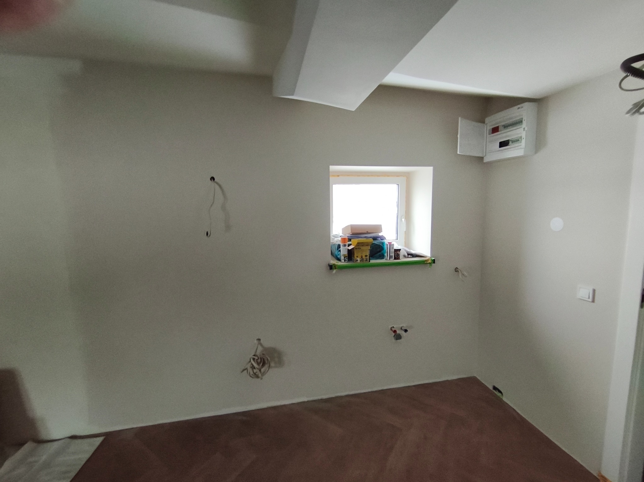 Wnętrze w trakcie remontu: świeżo pomalowane ściany, brązowa podłoga, okno z narzędziami, otwarta skrzynka elektryczna. Widoczne kable i instalacje.