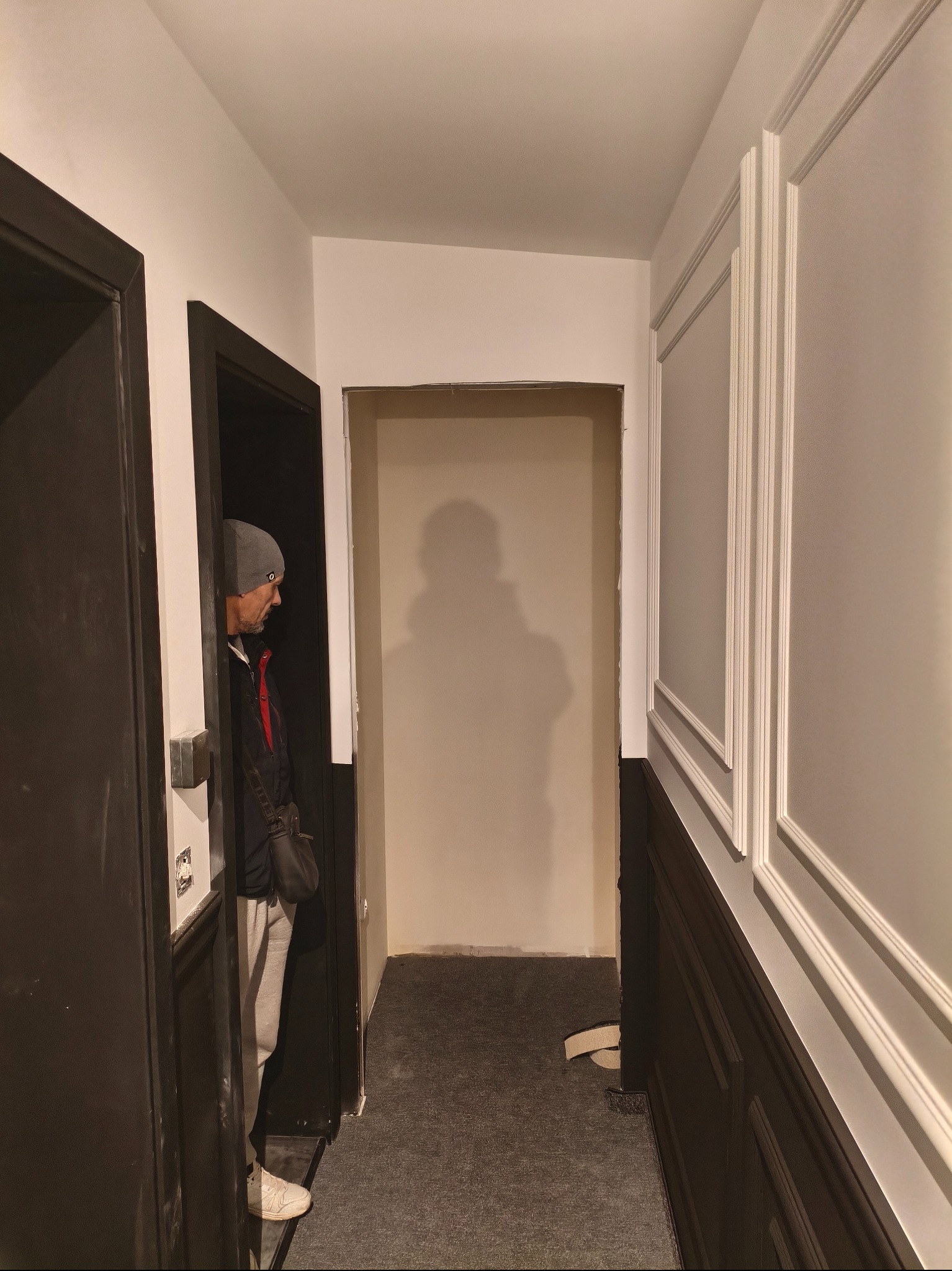 Korytarz po remoncie z panelami ściennymi i otwartym przejściem. Widoczny mężczyzna w drzwiach. Ciemne panele dolne kontrastują z białymi górnymi i dekoracyjnymi listwami.