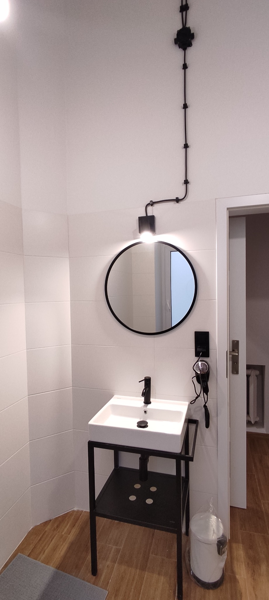 Nowoczesna łazienka z białą umywalką na czarnym stelażu, okrągłym lustrem z podświetleniem LED, czarną armaturą i nietypowym, natynkowym oświetleniem sufitowym.