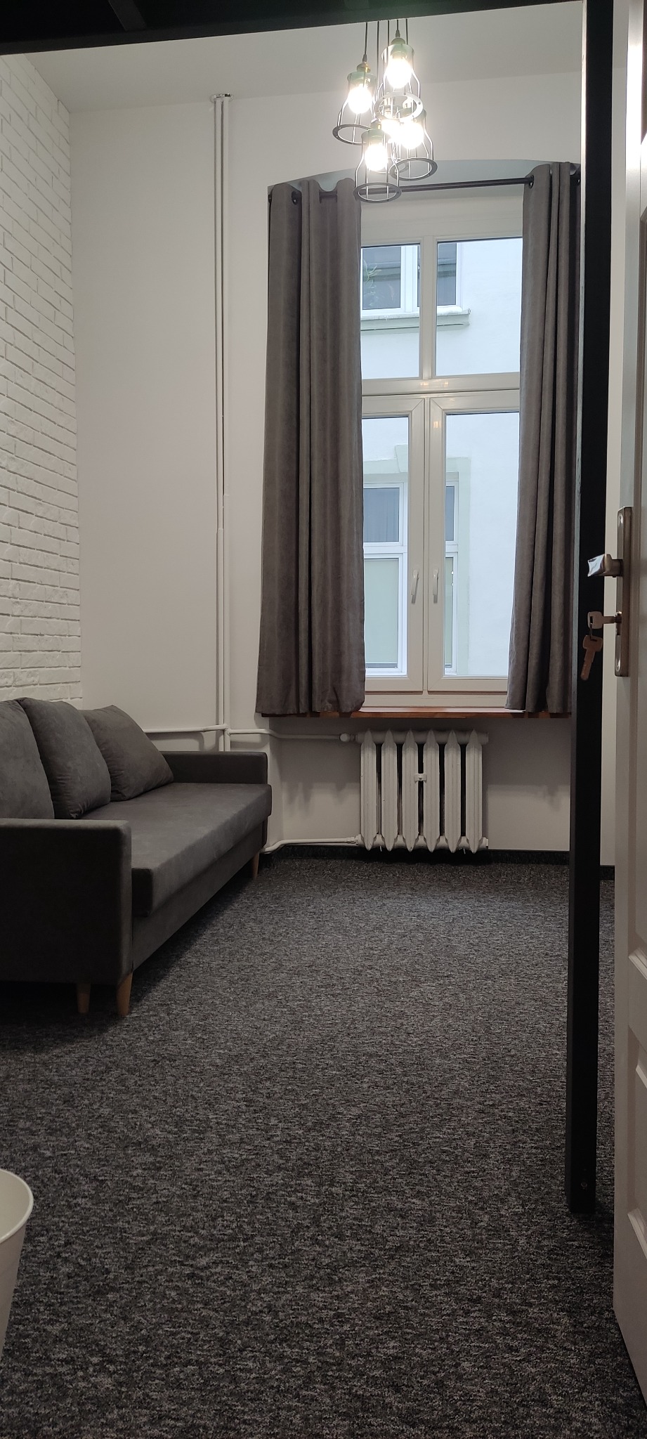 Jasne wnętrze pokoju z szarą sofą, grzejnikiem pod oknem z szarymi zasłonami i lampą sufitową w stylu industrialnym.