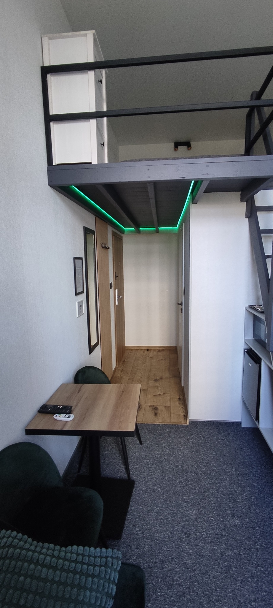 Funkcjonalne wnętrze z antresolą, oświetleniem LED, drewnianą podłogą i minimalistycznym wyposażeniem w odcieniach szarości i zieleni.