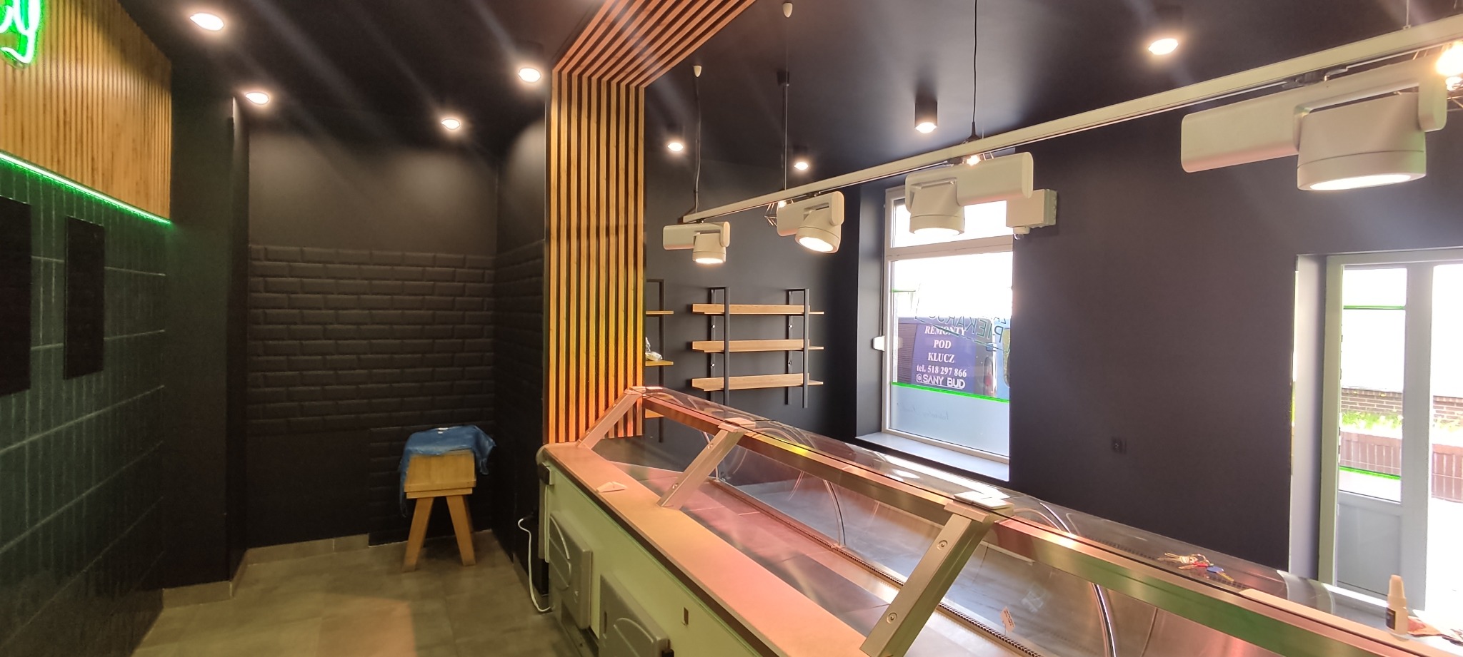 Wykończone wnętrze piekarni z czarnymi ścianami, drewnianymi lamelami, oświetleniem punktowym i chłodniczą witryną ekspozycyjną.