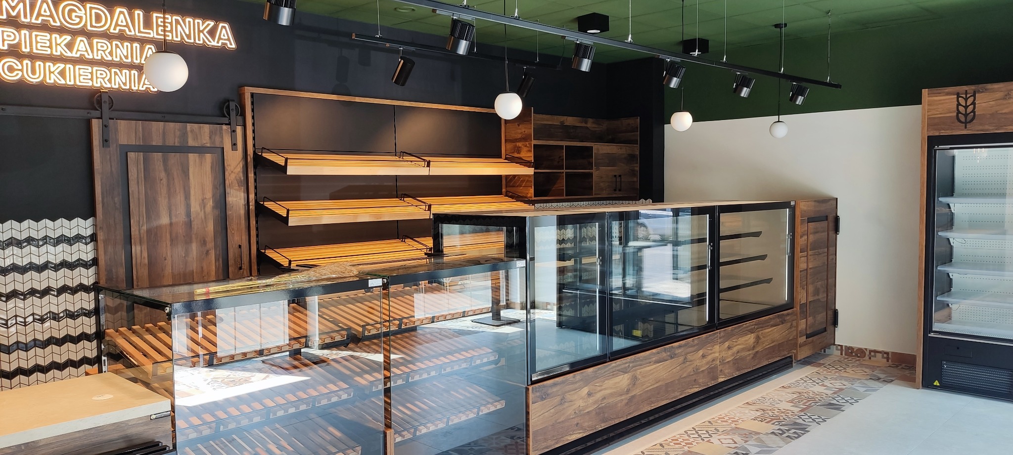 Nowoczesne wnętrze piekarni-cukierni z drewnianymi regałami, szklanymi witrynami chłodniczymi i dekoracyjną ścianą z mozaiką.