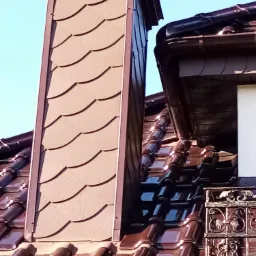 Brązowy komin obłożony łuskami dachowymi, dopasowany kolorystycznie do ceramicznej dachówki i rynny, na tle błękitnego nieba.