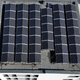 Widok z góry na instalację paneli słonecznych na płaskim dachu budynku z białymi ścianami, obok zaparkowany biały bus.